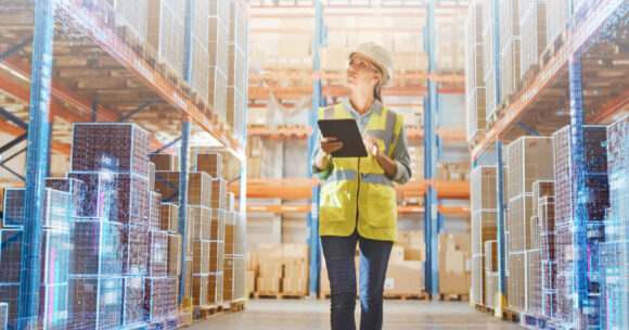 woman holding tablet looking up at shelves full of boxes in a large warehouse
